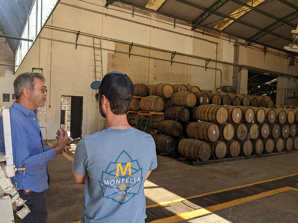 Inside a warehouse, along one side of the wall lines stacks of barrels, and two people discussing the barrels
